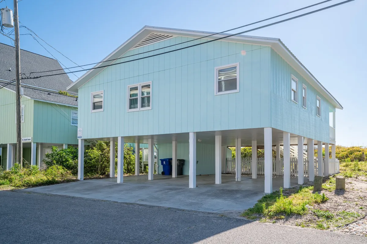 This image shows the exterior of the Casalona beach cottage, with its classic single-level structure elevated above the ground, surrounded by lush greenery. The inviting front porch features rocking chairs, perfect for sunset conversations, while the underneath space suggests easy beach access and room for family gatherings or barbecues.