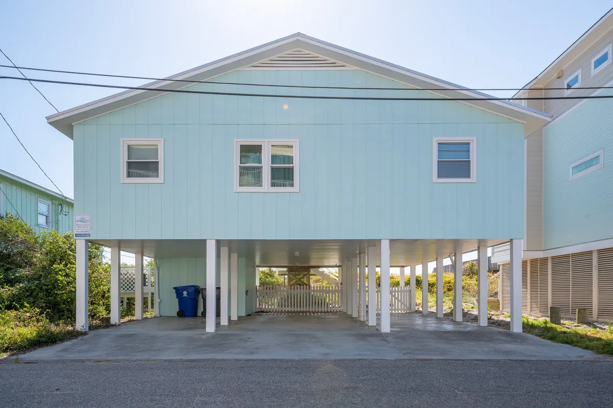 The exterior of the beach house features a light blue facade, spacious front porch, and the shaded area underneath the house. Sunset light softens the scene, highlighting spaces for gathering, relaxing, or barbecuing with friends and family.