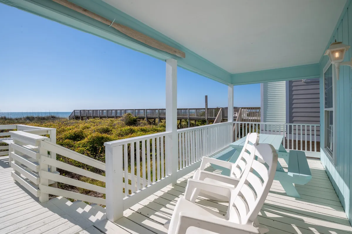 An inviting porch with comfortable deck seating and an outdoor table overlooks a sweeping ocean view. Gentle sunlight bathes the space, highlighting the proximity to the beach and offering a serene atmosphere perfect for lounging, dining, or enjoying morning coffee.