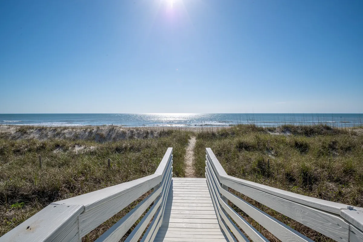 A private wooden boardwalk extends from the property toward the sandy beach, with sunlight glistening on the ocean beyond. The scene captures the seamless connection from porch to beach, emphasizing the easy access and allure of spontaneous outings.