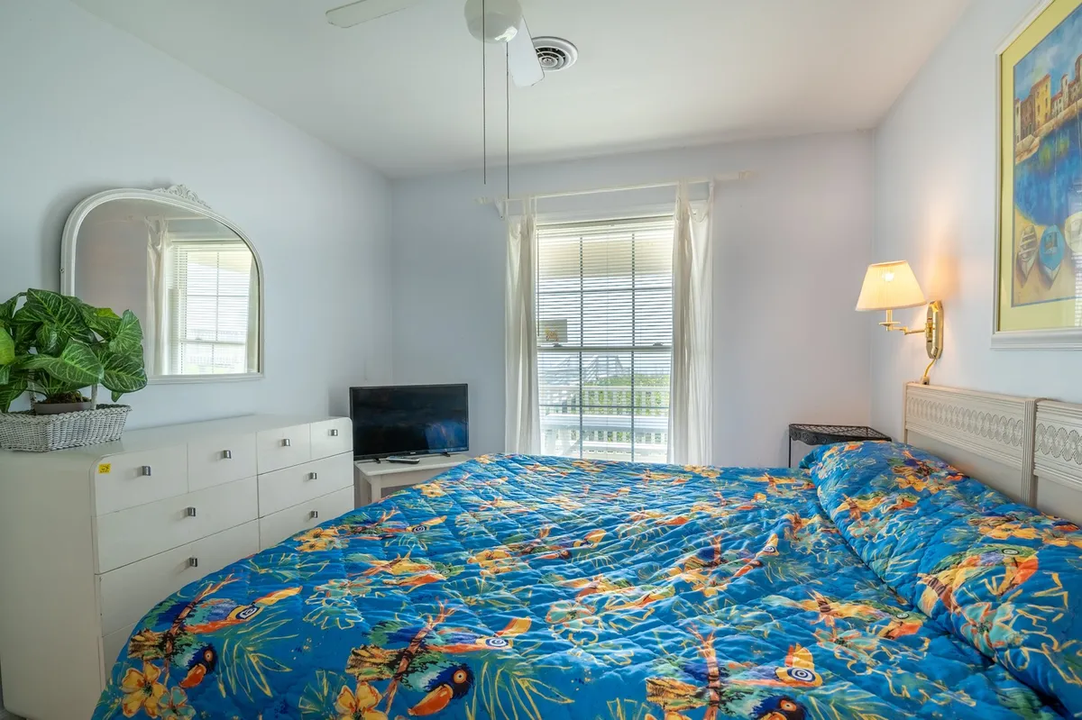 A bright bedroom with a colorful bedspread, large window letting in natural light, a plant, mirror, and TV. The bed is neatly made and exudes a fresh, welcoming feel, suggesting sealed linens and thoughtful touches.
