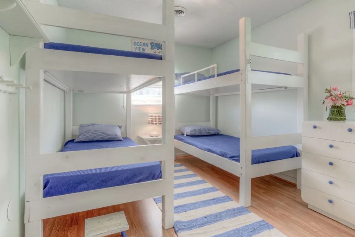 A playful bedroom with bunk beds, a striped rug, a white dresser, and a flower arrangement. The space suggests a welcoming environment for children or friends, encouraging both rest and fun activities.