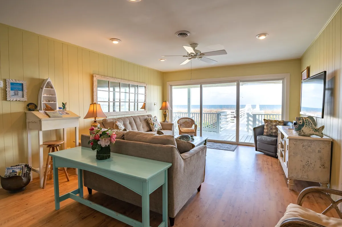 An inviting living area featuring large sliding glass doors that open to the deck, a comfortable sofa, a decorative table, and soft lighting from floor lamps. The room feels open, airy, and connected to the outdoors, with hints of ocean views and a layout perfect for entertaining or relaxing.