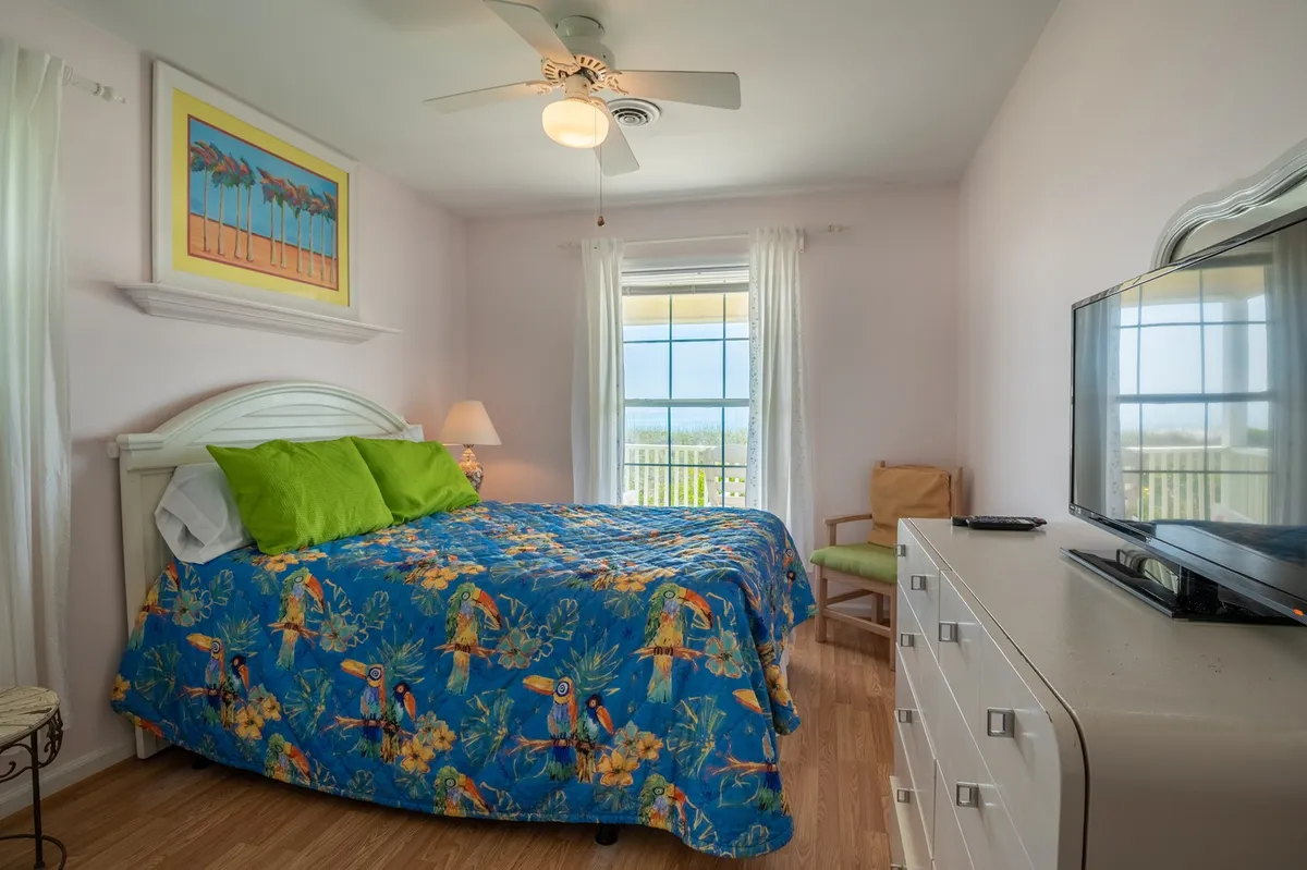 A vibrant bedroom featuring a colorful bedspread, a large window letting in natural light, a TV on a wooden dresser, and cozy wooden furniture. The room feels cheerful and welcoming, ideal for both relaxation and entertainment.