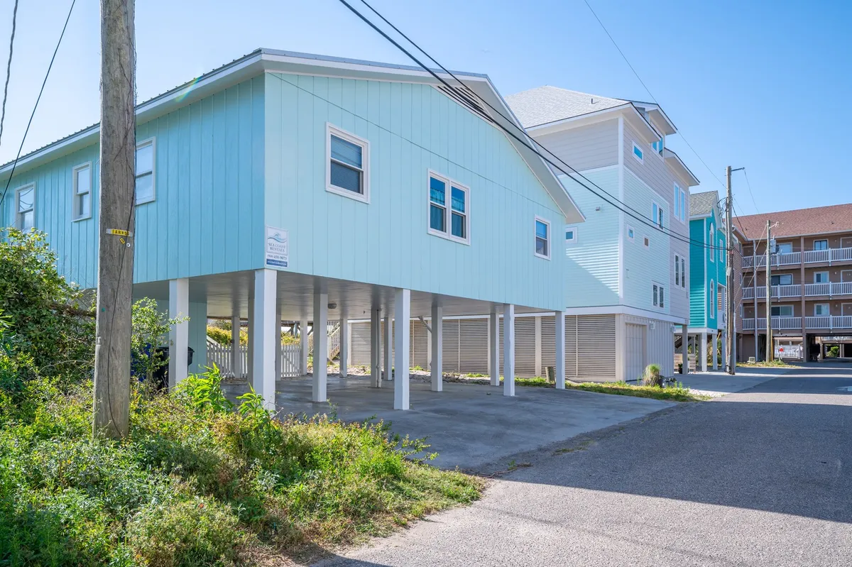 A relaxed exterior shot of the light blue beach house framed by green foliage and a concrete driveway, with easy access encouraging outdoor gatherings and evening strolls to the nearby beach.