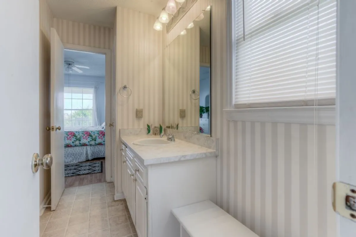 A serene Jack & Jill bathroom with a double sink vanity, mirror, striped wallpaper, and a door leading to a bedroom. The space is bright and functional, designed for shared yet private use.
