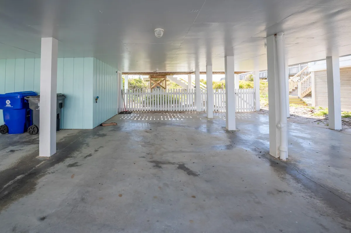 This image captures the area beneath the beach cottage, complete with sturdy support columns, a blue garbage bin, and a white picket fence. This space hints at practical seaside living—storing beach gear, hosting casual outdoor gatherings, and the functional aspects of a classic Carolina cottage.
