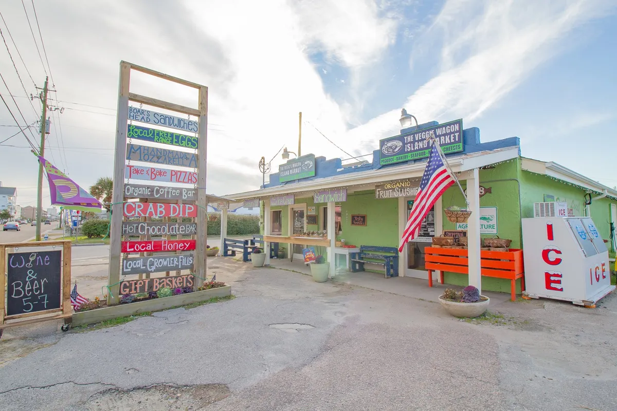 A colorful, inviting scene outside a local market or café, with vibrant signage, fresh produce displays, American flag, and outdoor seating where people enjoy coffee or socialize.
