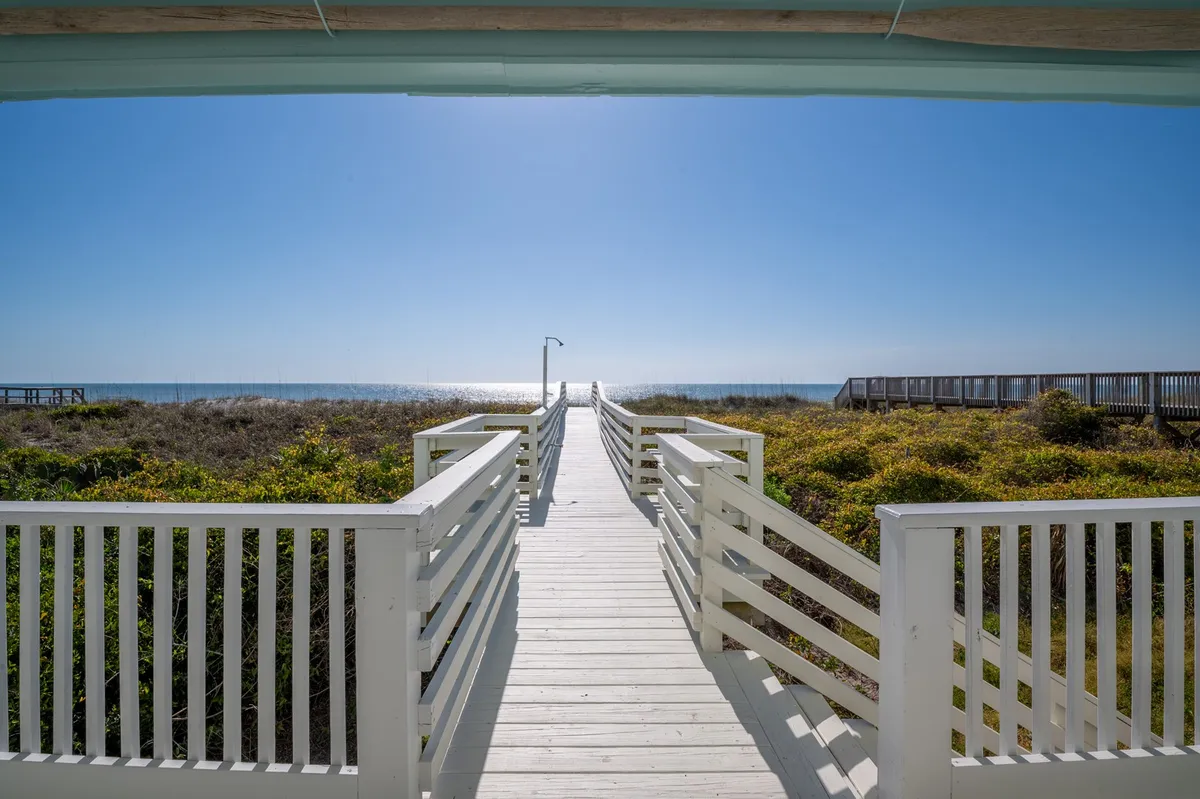 A serene exterior shot of a wooden boardwalk stretching out toward the ocean, surrounded by natural coastal vegetation, with gentle waves visible in the distance. It evokes the feeling of peaceful strolls and easy access between the house and the shore.