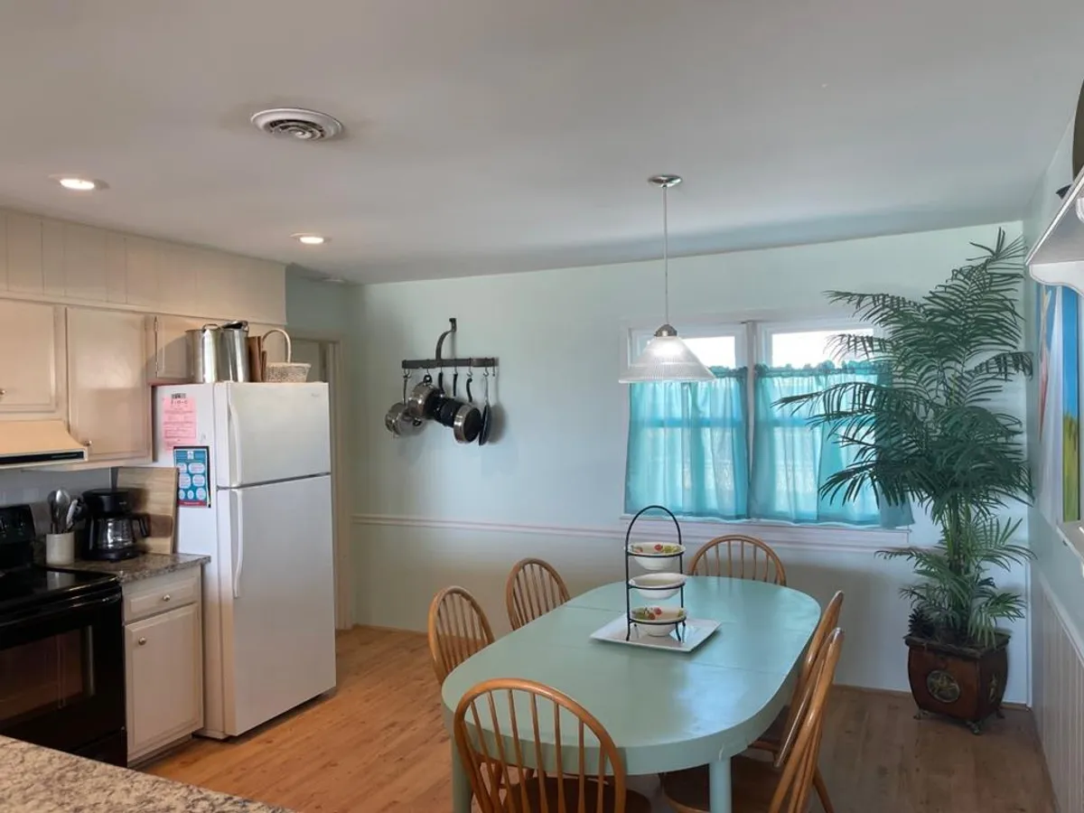 A warm and welcoming dining area that seamlessly connects to the kitchen. The dining table is set for a family meal, with a kitchen counter visible in the background, potted plants adding a touch of vibrancy, and sunlight streaming through windows with curtains. The arrangement encourages shared meals and conversation.