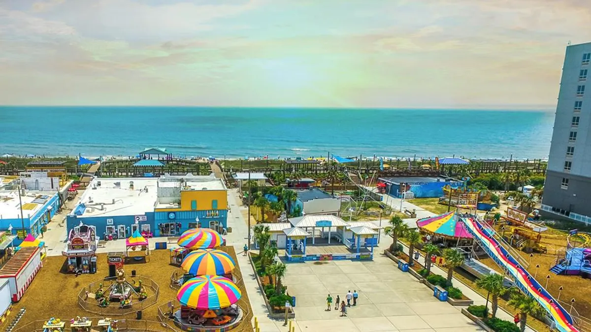 A lively boardwalk scene featuring colorful umbrellas, amusement park rides, the ocean in the background, and people walking and enjoying the atmosphere. The image conveys the energy and fun of the Carolina Beach Boardwalk during summertime events.