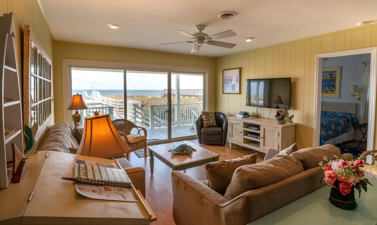 A bright, open living area featuring large windows with an expansive ocean view, a cozy seating arrangement with a comfortable sofa and armchairs, a decorative lamp, a coffee table, and a television. The space feels casual yet inviting, with natural light flooding the room and a welcoming atmosphere for gathering or relaxing.